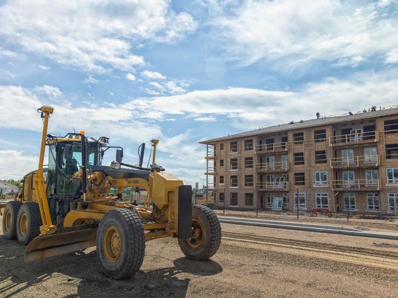 A grader works on a city road in front of a multi-story building under construction.