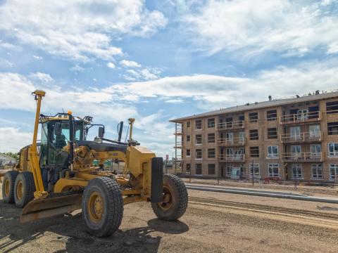 A grader works on a city road in front of a multi-story building under construction.
